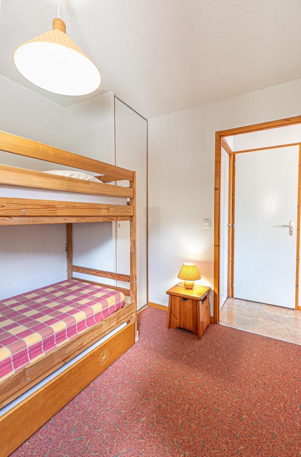 Un lit superposé avec un matelas à carreaux, un petit tabouret en bois sur lequel est posée une lampe, et une porte visible au fond.