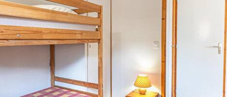 A bunk bed with a checkered mattress, a small wooden stool with a lamp, and a door in the background.