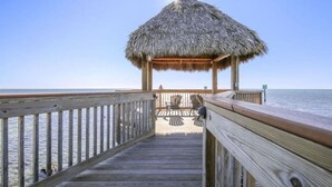 Une jetée en bois menant à une structure au toit de chaume, dotée d’un espace extérieur aménagé pour s’asseoir.