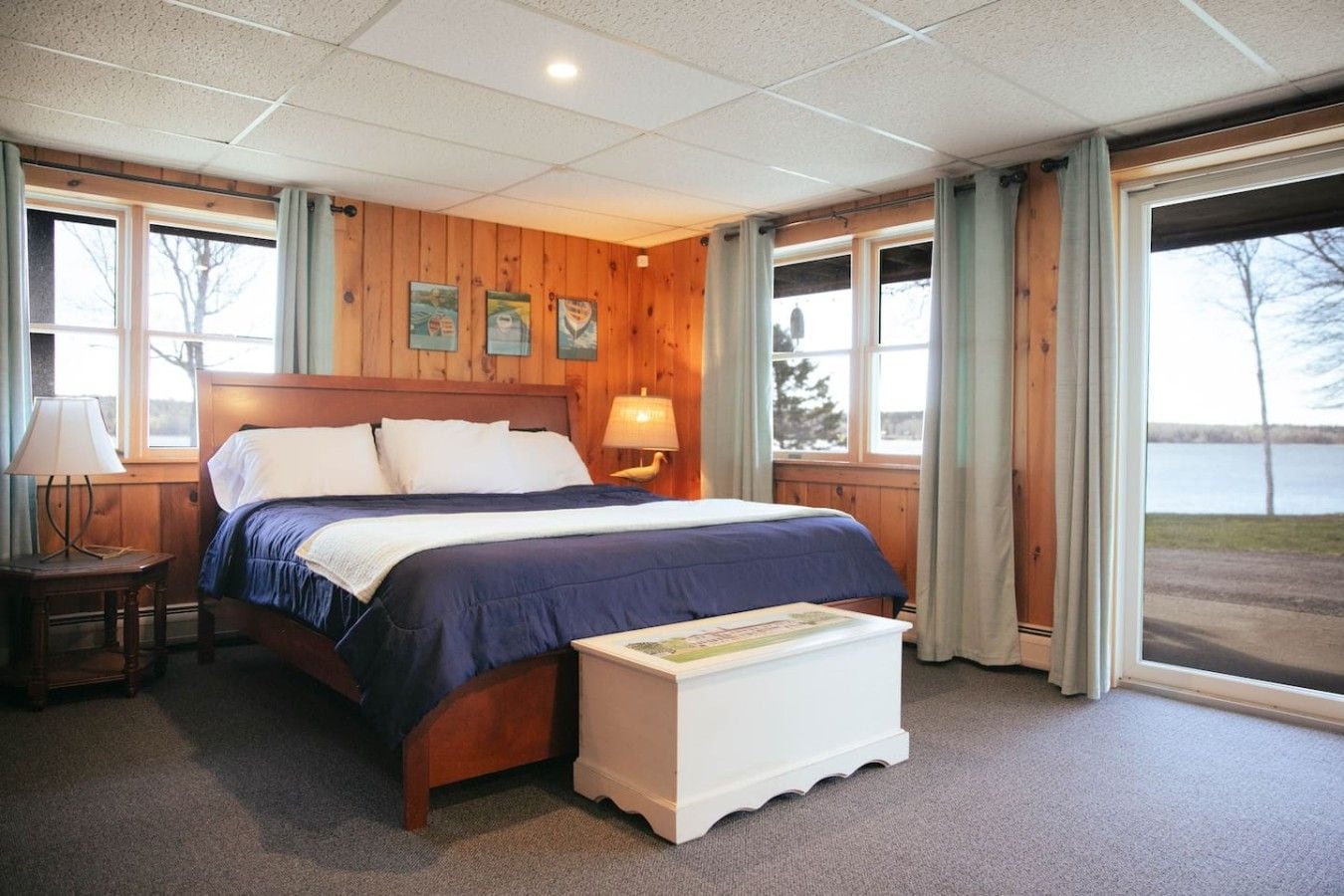A hotel room with a wooden paneled wall, a large bed with a blue bedspread, a bedside table with a lamp, and a view of a body of water through the window.