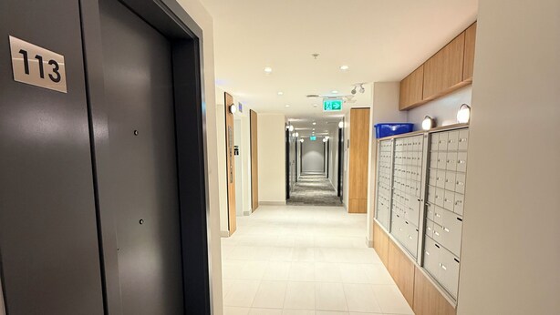 A hotel hallway with a carpeted floor, a row of lockers on the right, and a door labeled '113' on the left. - Modern Condo Steps From SkyTrain-Walkable Location + Amazing Building Amenities (Surrey)