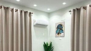 Une chambre d’hôtel avec un lit, des rideaux, un appareil de climatisation, une plante en pot et un tableau accroché au mur.
