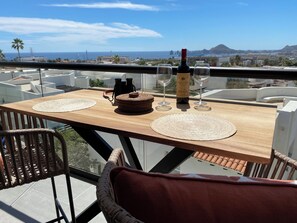 Ein Holztisch mit einer Weinflasche, zwei Gläsern und einem Fernglas auf einem Balkon mit Blick auf eine Stadtlandschaft und das Meer.