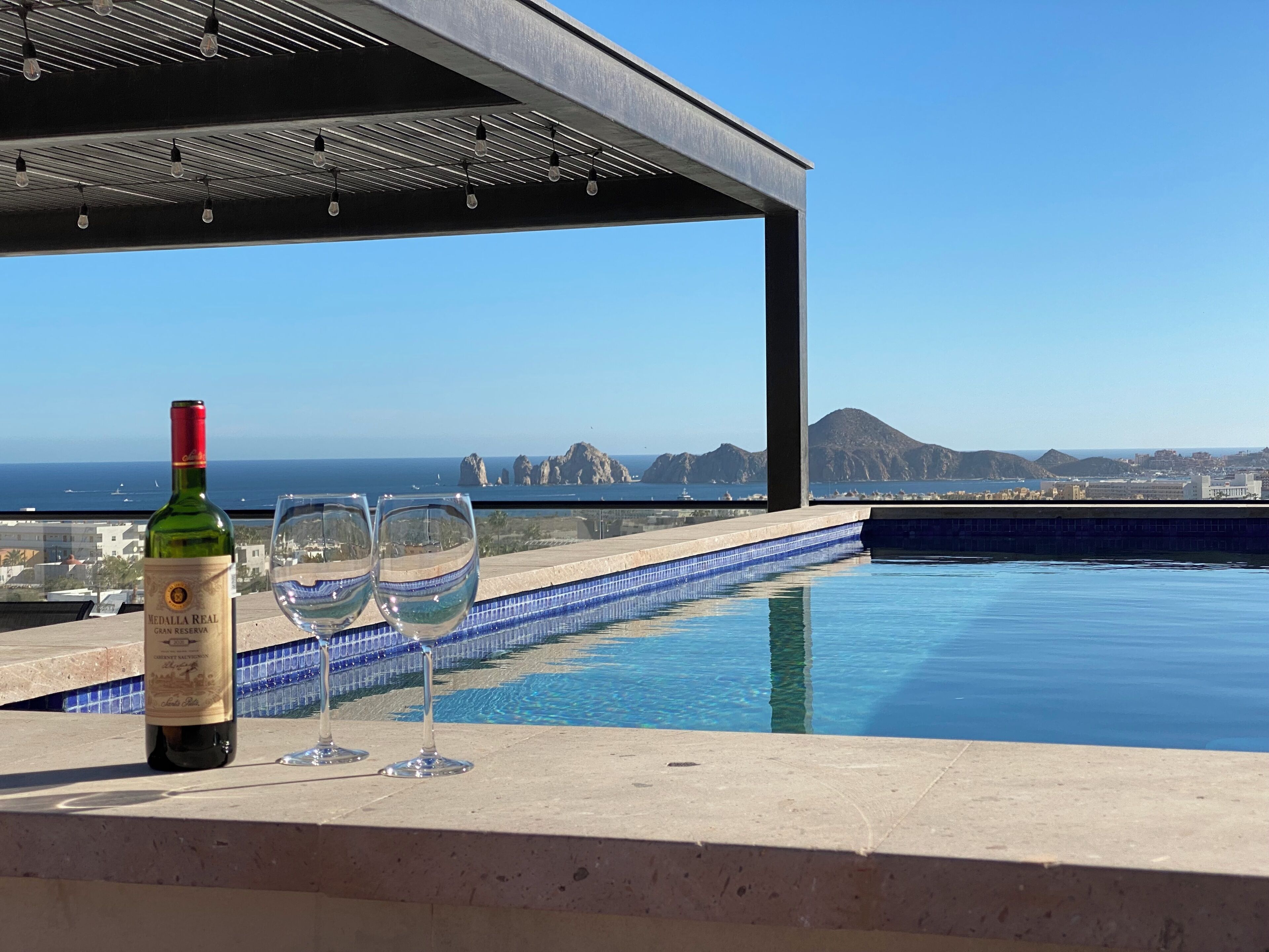 A poolside setting with a bottle of wine and glasses, overlooking a coastal landscape with buildings and a mountain.