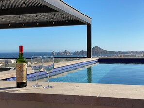 Ein Poolbereich mit einer Weinflasche und Gläsern, mit Blick auf eine Küstenlandschaft mit Gebäuden und einem Berg.