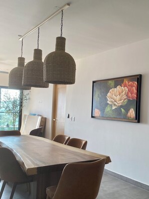 Un coin repas avec une table et des chaises en bois, un luminaire suspendu en fibres tressées et un tableau floral encadré accroché au mur.