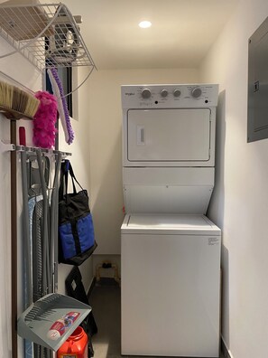 Une buanderie équipée d’un lave-linge et d’un sèche-linge Whirlpool blancs, d’un étendoir en métal avec des produits de nettoyage et d’une bouteille de gaz Kirkland rouge.