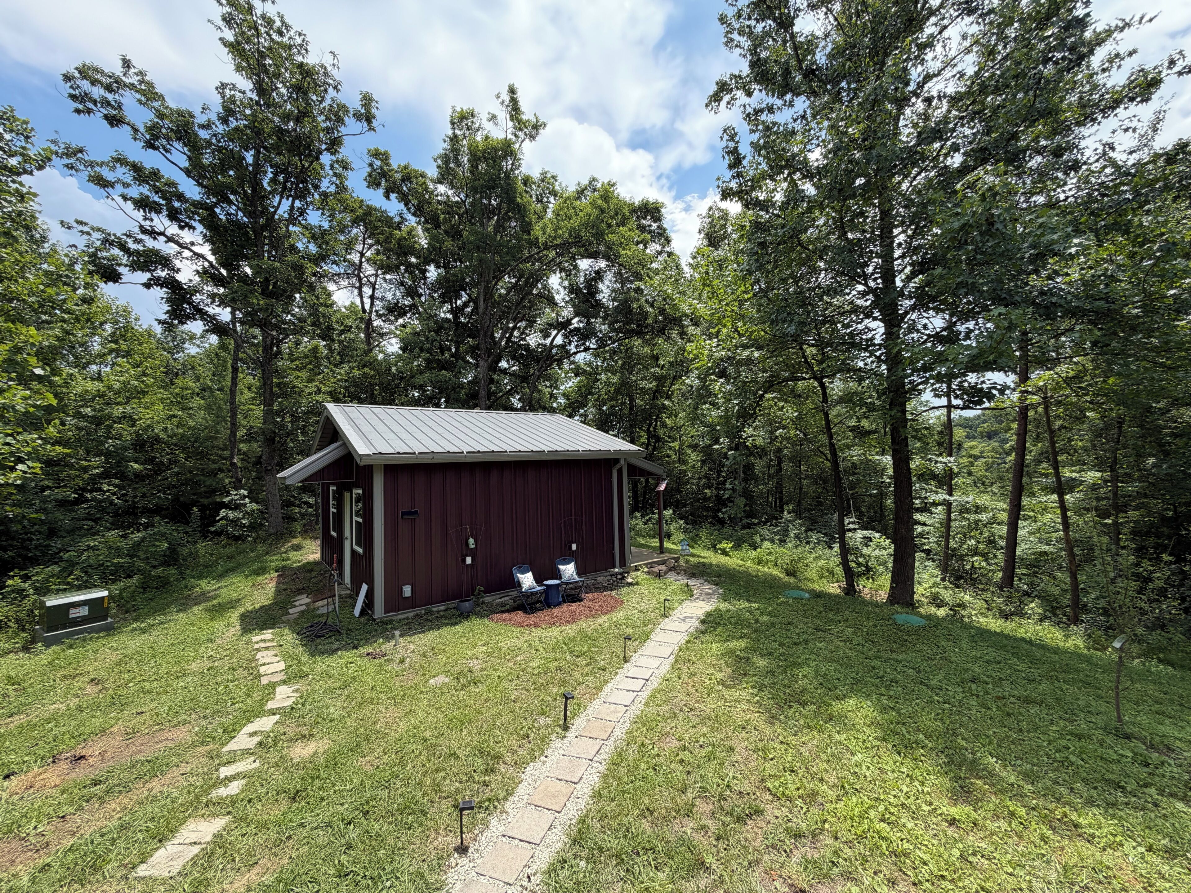 A small, single-story wooden cabin with a metal roof, surrounded by a grassy area and dense trees.