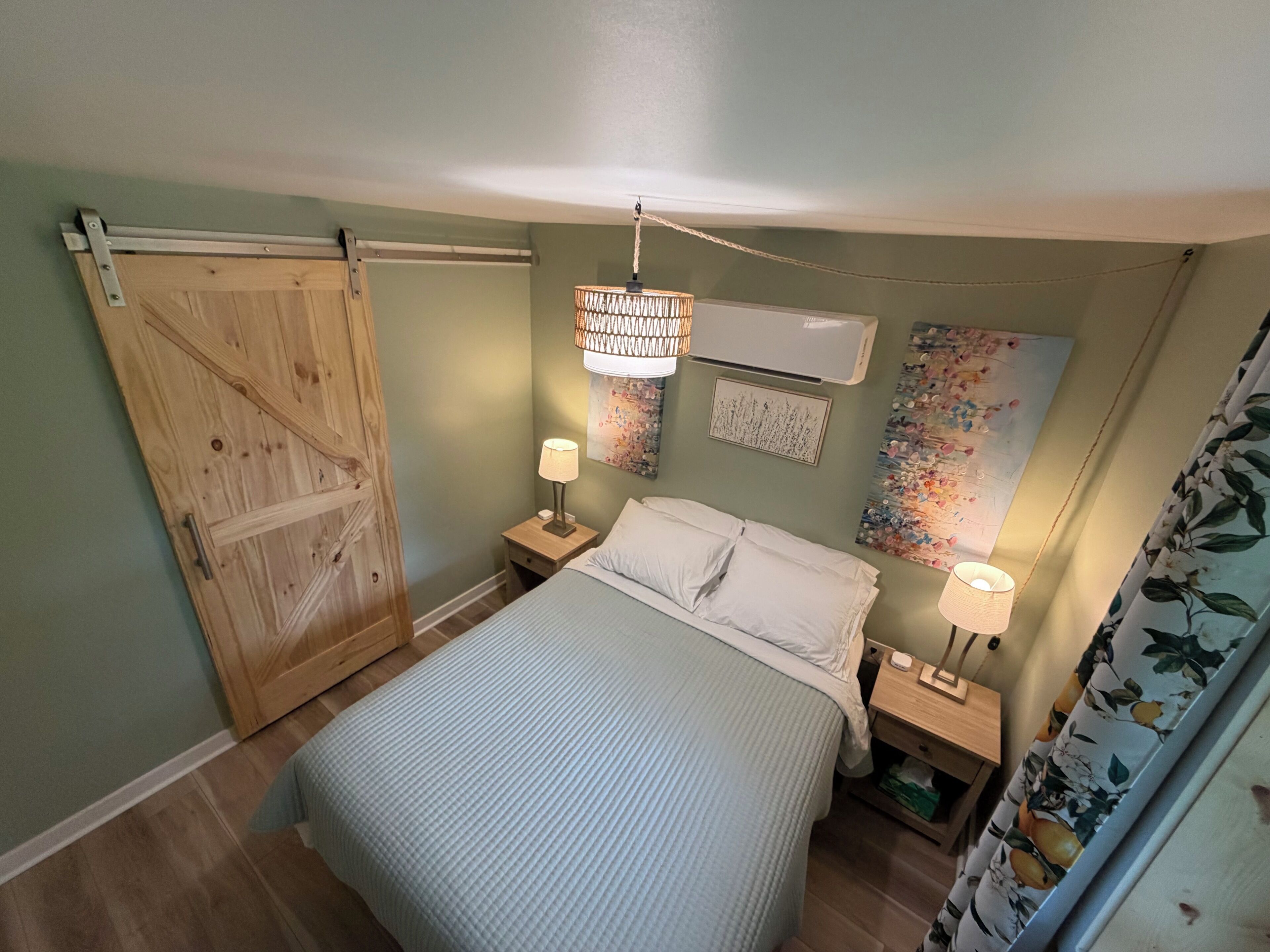 A bedroom with a bed, two bedside tables, a hanging light fixture, an air conditioning unit, and a door.