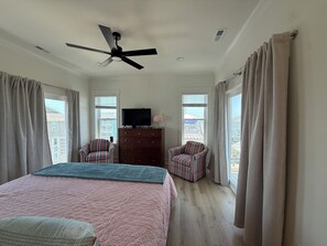 A bedroom with a bed, two chairs, a TV on a dresser, a ceiling fan, and large windows with curtains. - New Luxury Beach Retreat  4 Bedrooms 3 King beds  Elevator  Steps to the Beach (Carolina Beach)