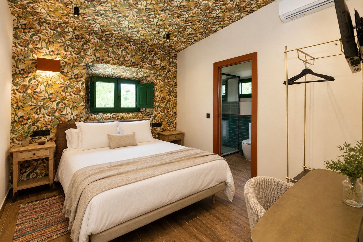 Quarto com papel de parede floral, cama com lençóis brancos, piso de madeira, uma pequena mesa com um vaso e luminária pendente.