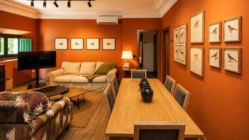 A living room with a sofa, a TV, a dining table with chairs, and framed pictures on the wall.