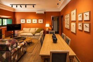 A living room with a sofa, a TV, a dining table with chairs, and framed pictures on the wall.