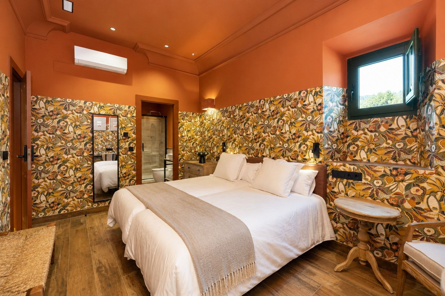 A bedroom with a large bed, a bedside table, a mirror, a window, and a bathroom with a toilet and sink.