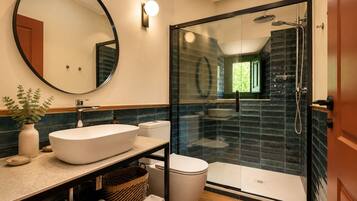 Un baño moderno con un espejo redondo, un lavabo grande, un inodoro y una zona de ducha con mampara de cristal.