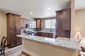 A kitchen with wooden cabinets, granite countertops, a microwave, and a window with blinds.