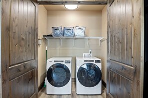 A laundry room with two front-loading washers and dryers, wooden doors, a shelf with bags, and a broom.