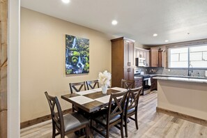 Un coin repas avec une table et des chaises en bois, un tableau accroché au mur et une cuisine en arrière-plan.