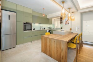 Une cuisine moderne dotée d’un four encastré, d’un coin repas avec une table en bois et des chaises jaunes, et d’un éclairage suspendu.