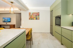 A modern kitchen with green cabinets, a wooden dining table, and a colorful wall art piece. - Lush 3-bed - Parking - 500Mbps - Riviera (Paleo Faliro)