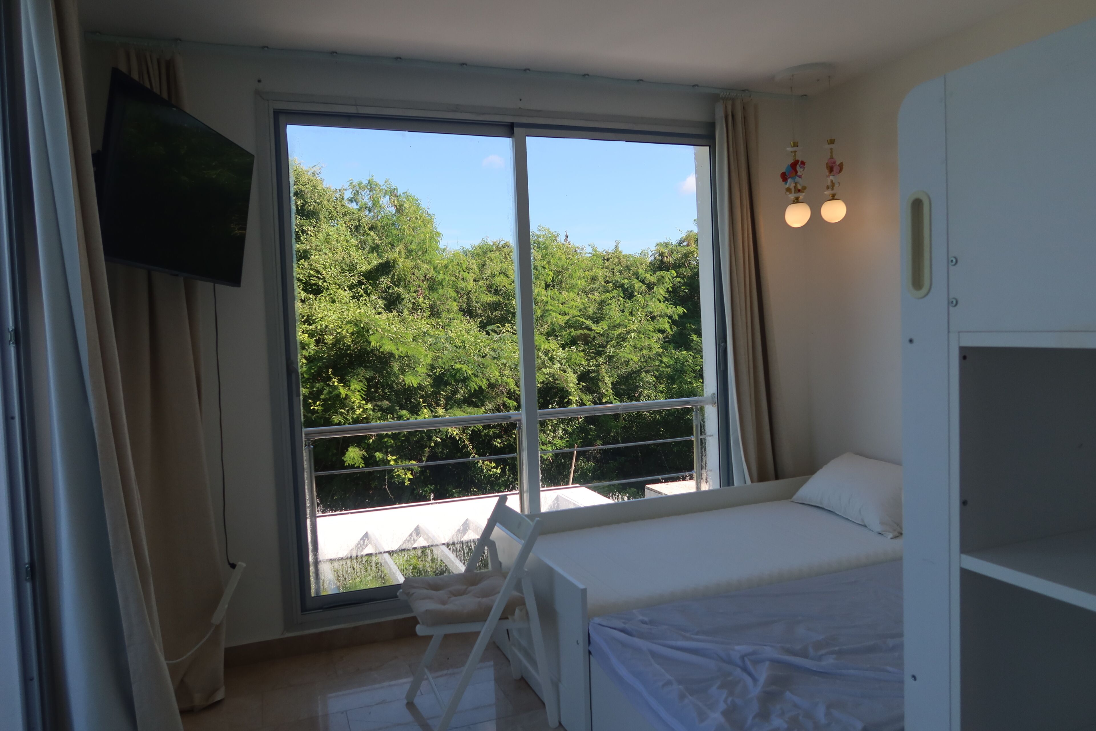A room with a bed, a TV mounted on the wall, a window with a view of trees, and a balcony.