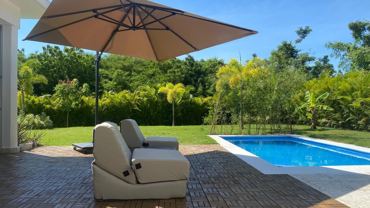 Et poolområde med en beige liggestol, en parasol og en swimmingpool omgivet af frodigt grønt.