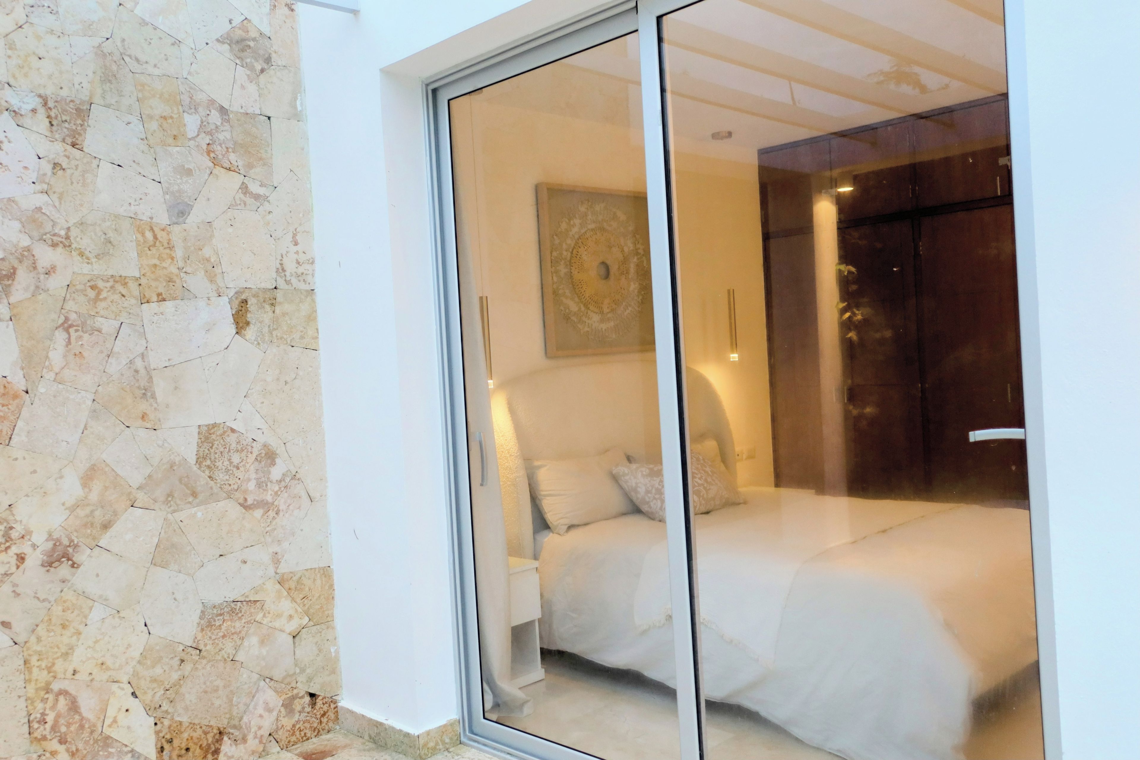 Une chambre avec une porte en verre donnant sur une salle de bain, un lit avec une literie blanche et une œuvre d’art accrochée au mur.