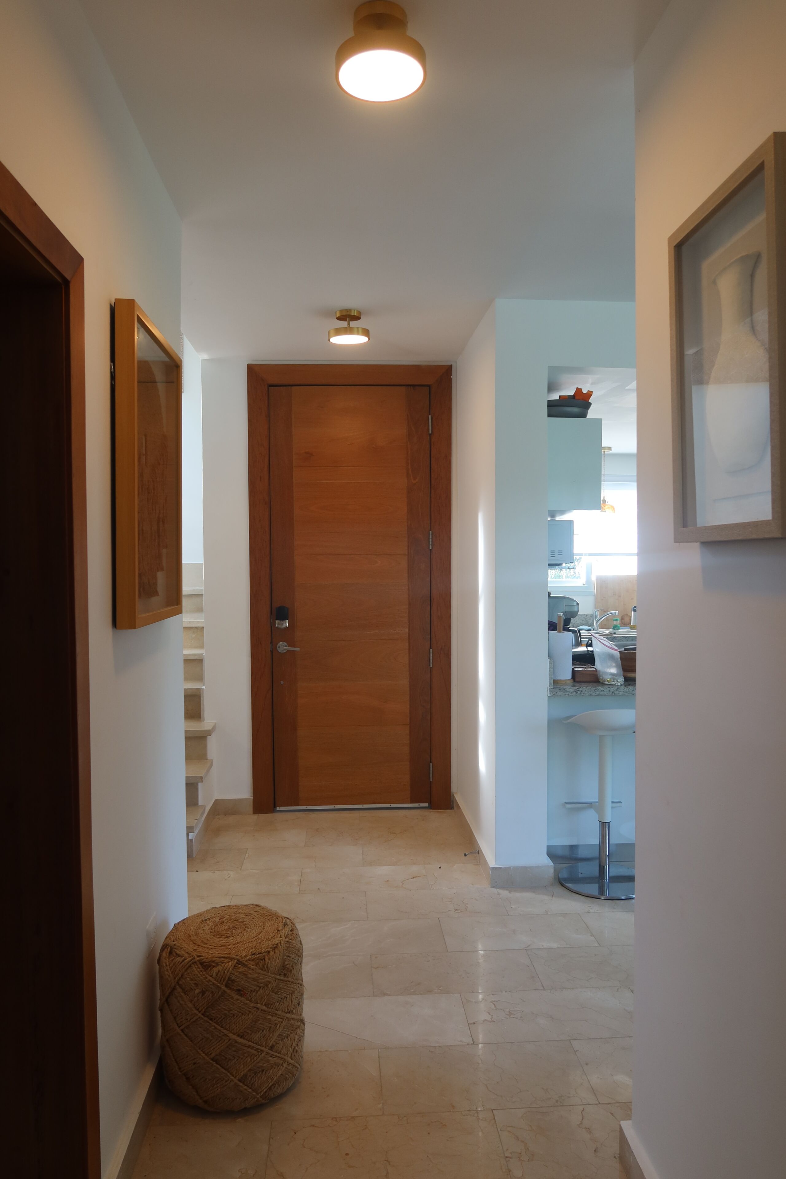 A hallway with a wooden door, a framed picture on the wall, a small round ottoman, and a kitchen area in the background.