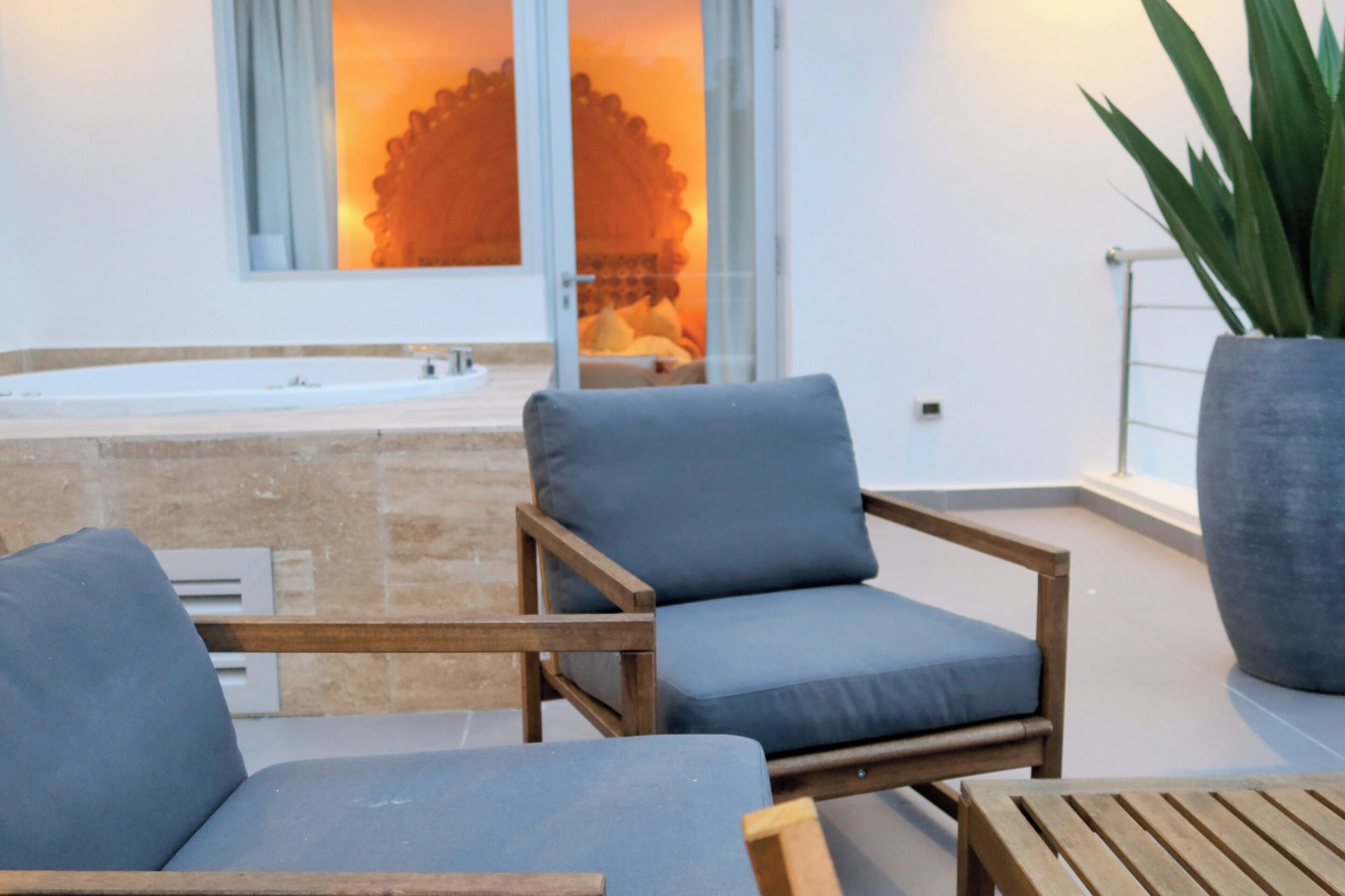 Ein moderner Außenbereich mit zwei Stühlen, einem Tisch, einer großen Pflanze und Blick auf ein Spa durch eine Glastür.