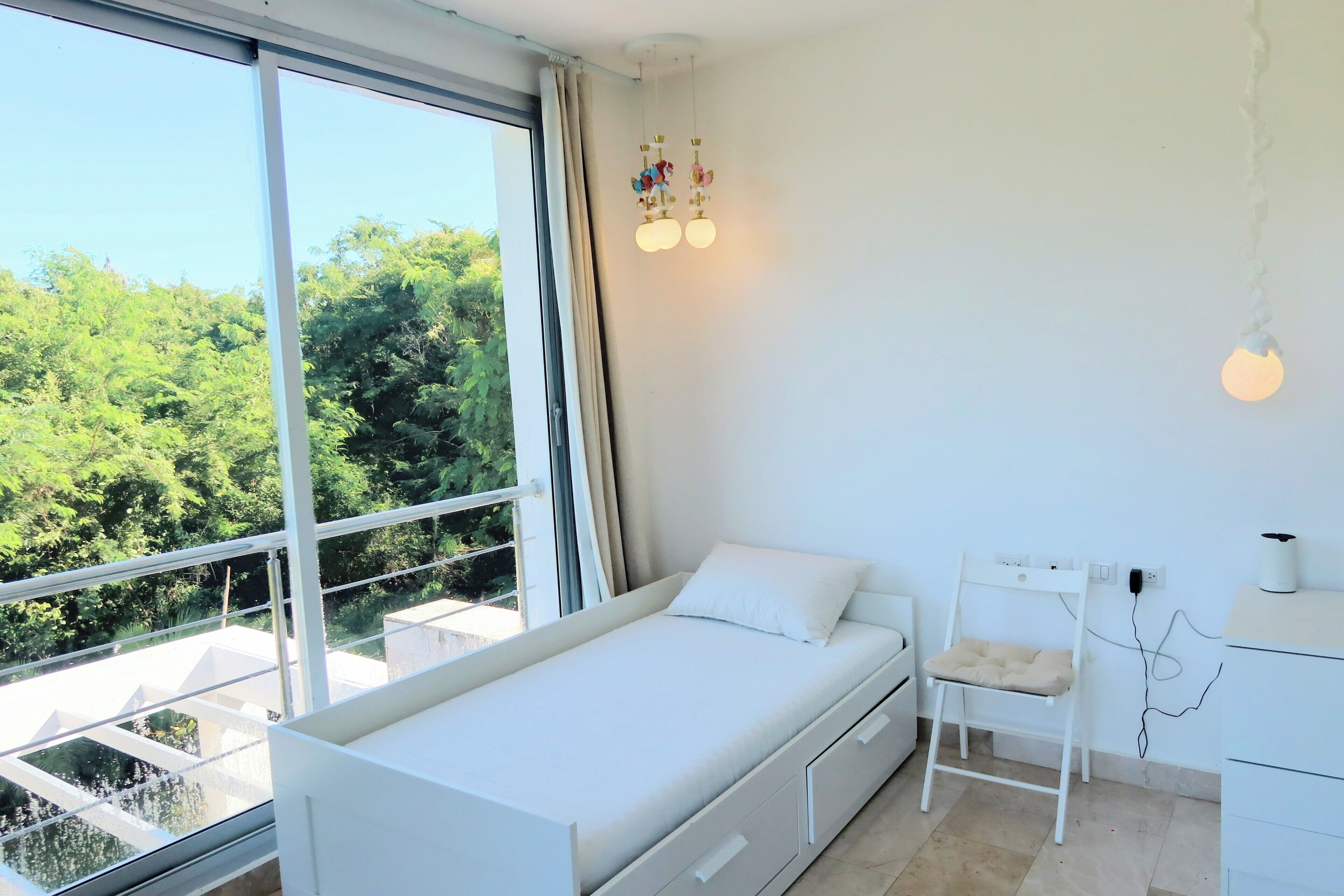 A bedroom with a bed, a desk, and a chair. There is a large window with a view of trees.