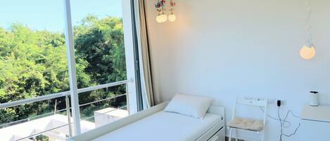 A bedroom with a bed, a desk, and a chair. There is a large window with a view of trees.