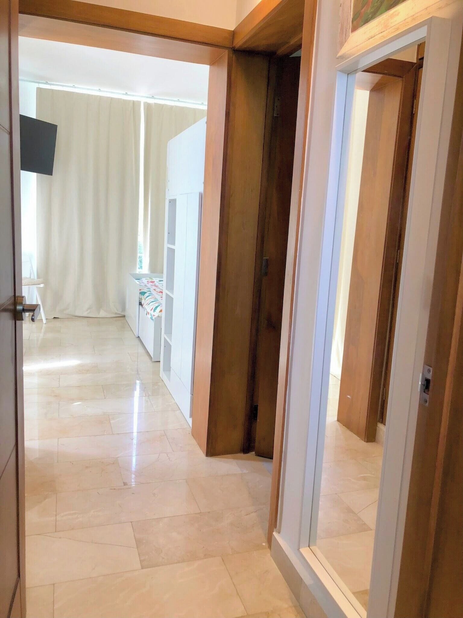 A corridor with a tiled floor leading to a room with a bed and a window.