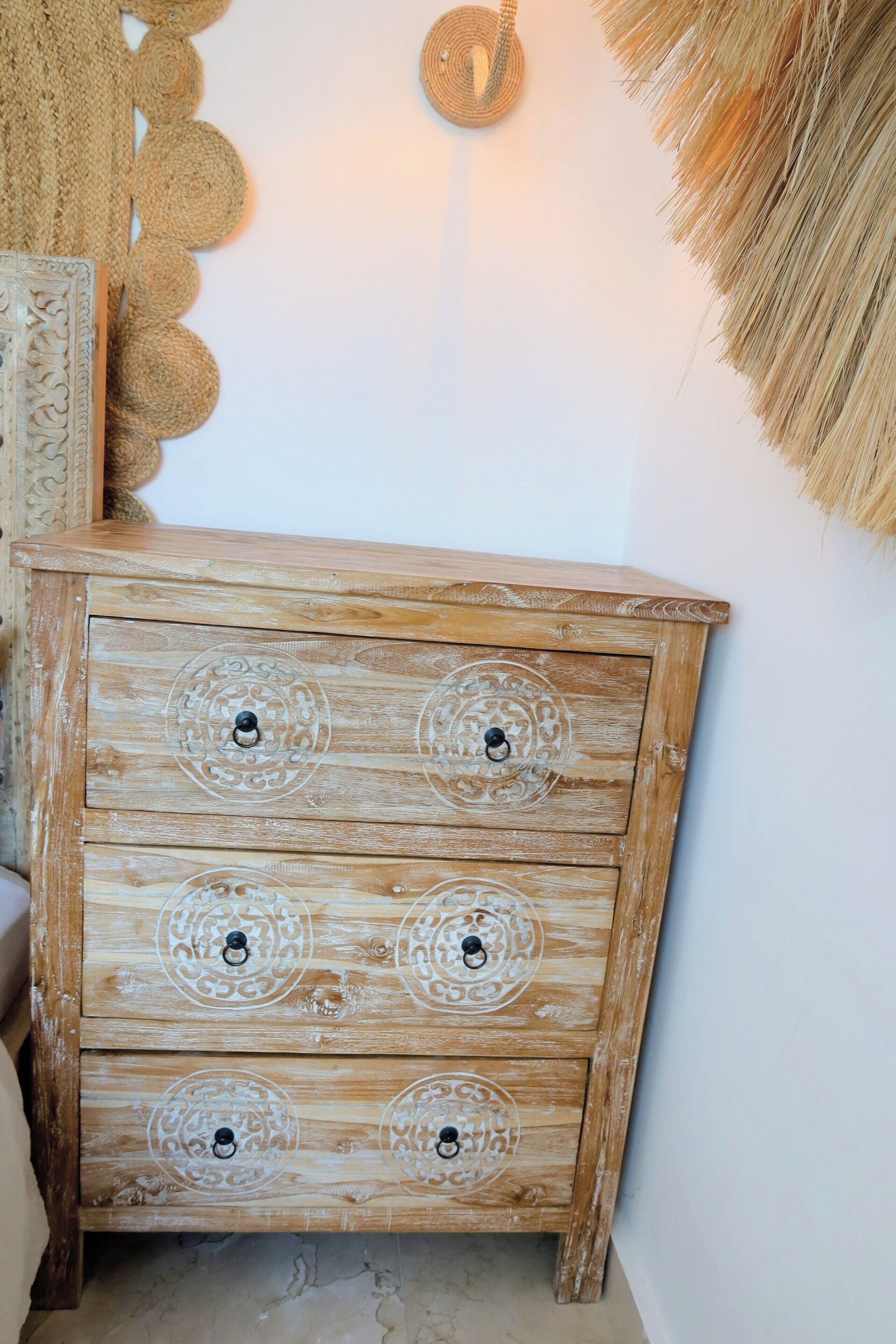 Een houten dressoir met versierde gravures en zwarte knoppen, geplaatst voor een muur met een geweven wanddecoratie en een structuurrijke wandversiering.