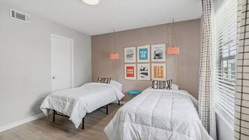 Un dormitorio con dos camas, una ventana con persianas, un conducto de ventilación en la pared y cuadros enmarcados.