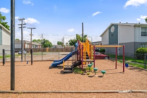 A playground with a slide and climbing structures, surrounded by a fence and residential buildings. - Spacious 3 Bedroom Apartment: Perfect for Families (Houston)