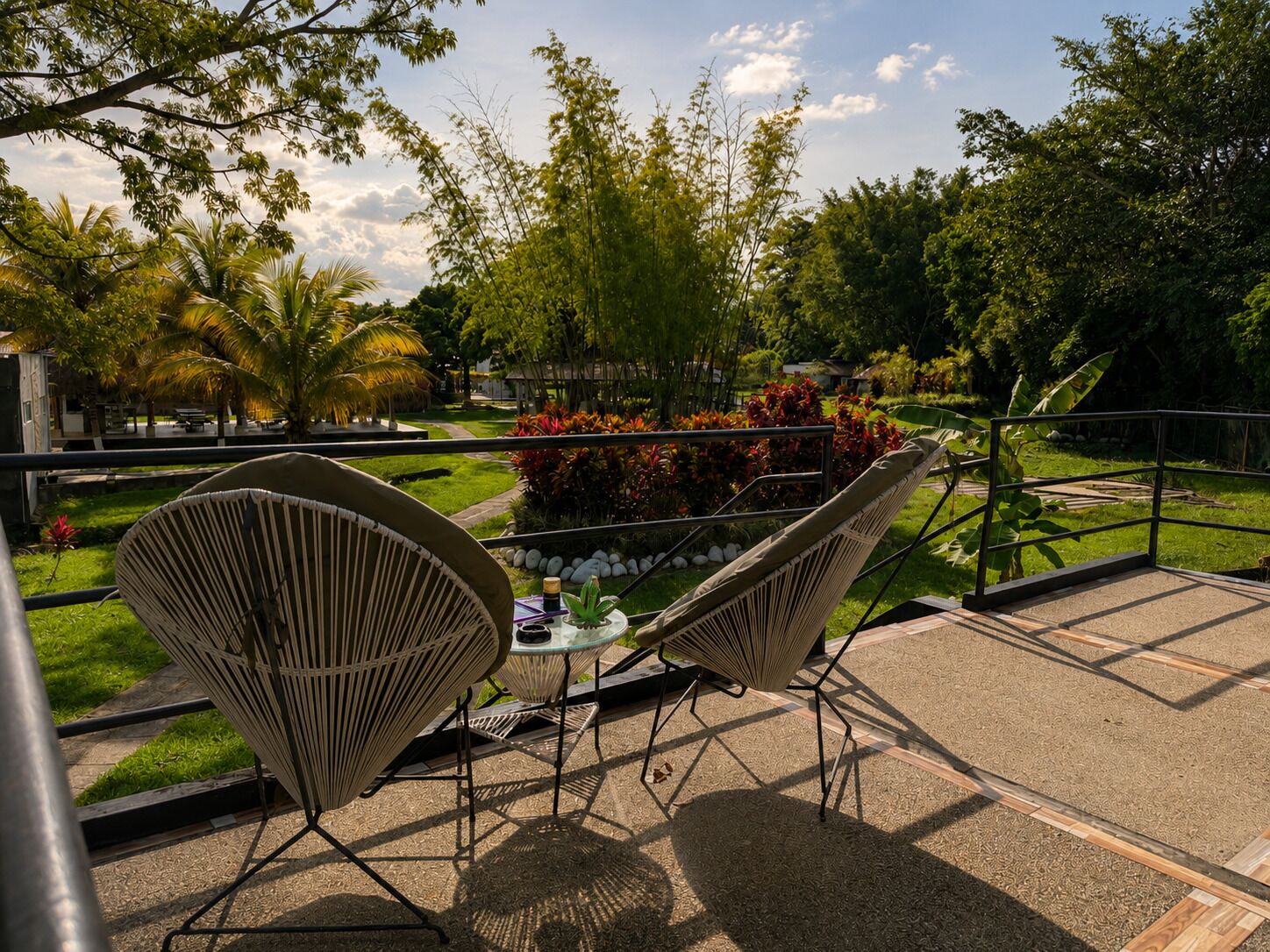 A terrace with two wicker chairs, a small table, and a view of a landscaped garden with various plants and trees.