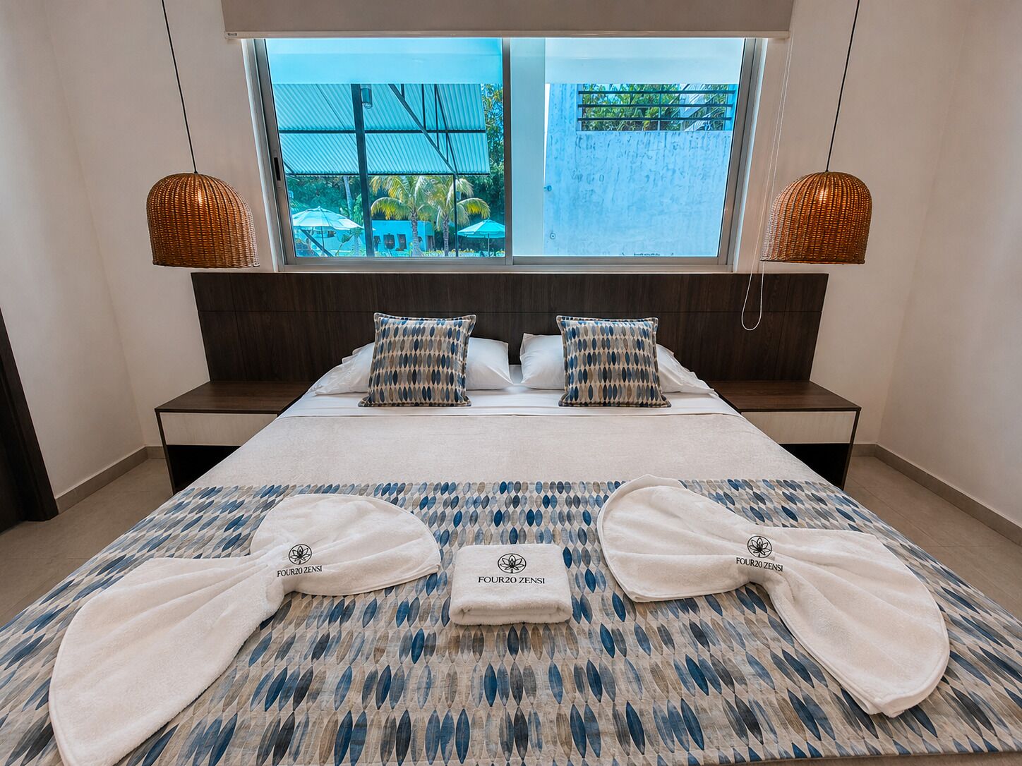 A hotel room with a bed, two bedside tables, a window with a view of the pool, and two hanging lamps.