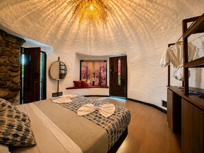 A cozy bedroom with a stone wall, a round mirror, a bed with a patterned bedspread, and a wooden bench by a window with a view of pink flowers. - Hotel Four20 Zensi - Meta (Lejanías)