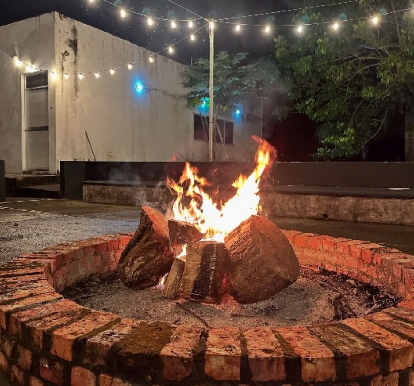 Un brasero circular con leña encendida, rodeado de un borde de ladrillo, con un edificio y luces colgantes de fondo.