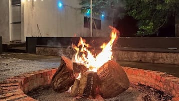 Un brasero circular con leña encendida, rodeado de un borde de ladrillo, con un edificio y luces colgantes de fondo.