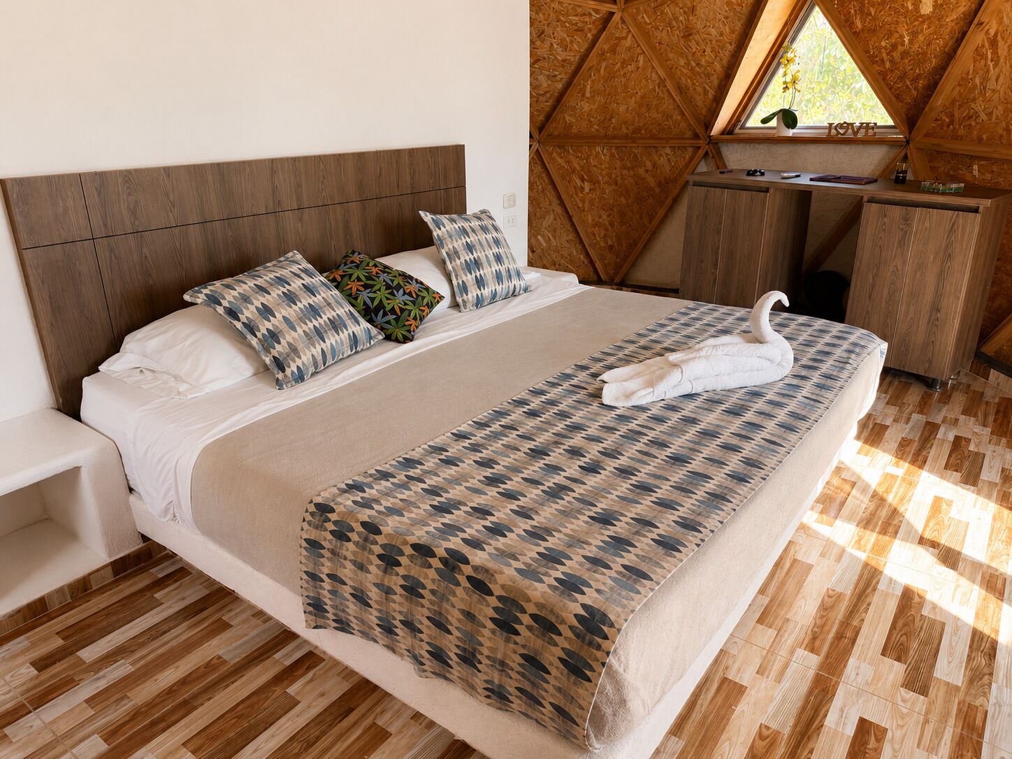 Un dormitorio con cabecera de madera, cama con ropa de cama estampada, toalla con forma de cisne, escritorio con un cartel que dice 'LOVE' y ventana con vistas a follaje.