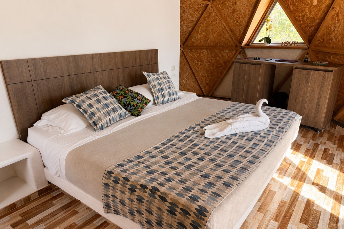 Una cama bien hecha con un almohadón con forma de cisne, un cabecero de madera y una ventana con vistas a la vegetación.