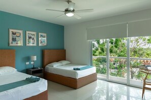 Ein Hotelzimmer mit zwei Betten, einem Deckenventilator, einem Nachttisch, einer Lampe und einem großen Fenster mit Blick auf Grünflächen.