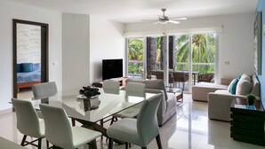 A modern living room with a glass dining table, comfortable seating, a flat-screen TV, and large windows offering a view of greenery. - Mareazul, Playa del Carmen, Oceanfront Condo, Paraiso Azul (Playa del Carmen)