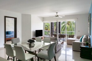 A modern living room with a glass dining table, comfortable seating, a flat-screen TV, and large windows offering a view of greenery. - Mareazul, Playa del Carmen, Oceanfront Condo, Paraiso Azul (Playa del Carmen)