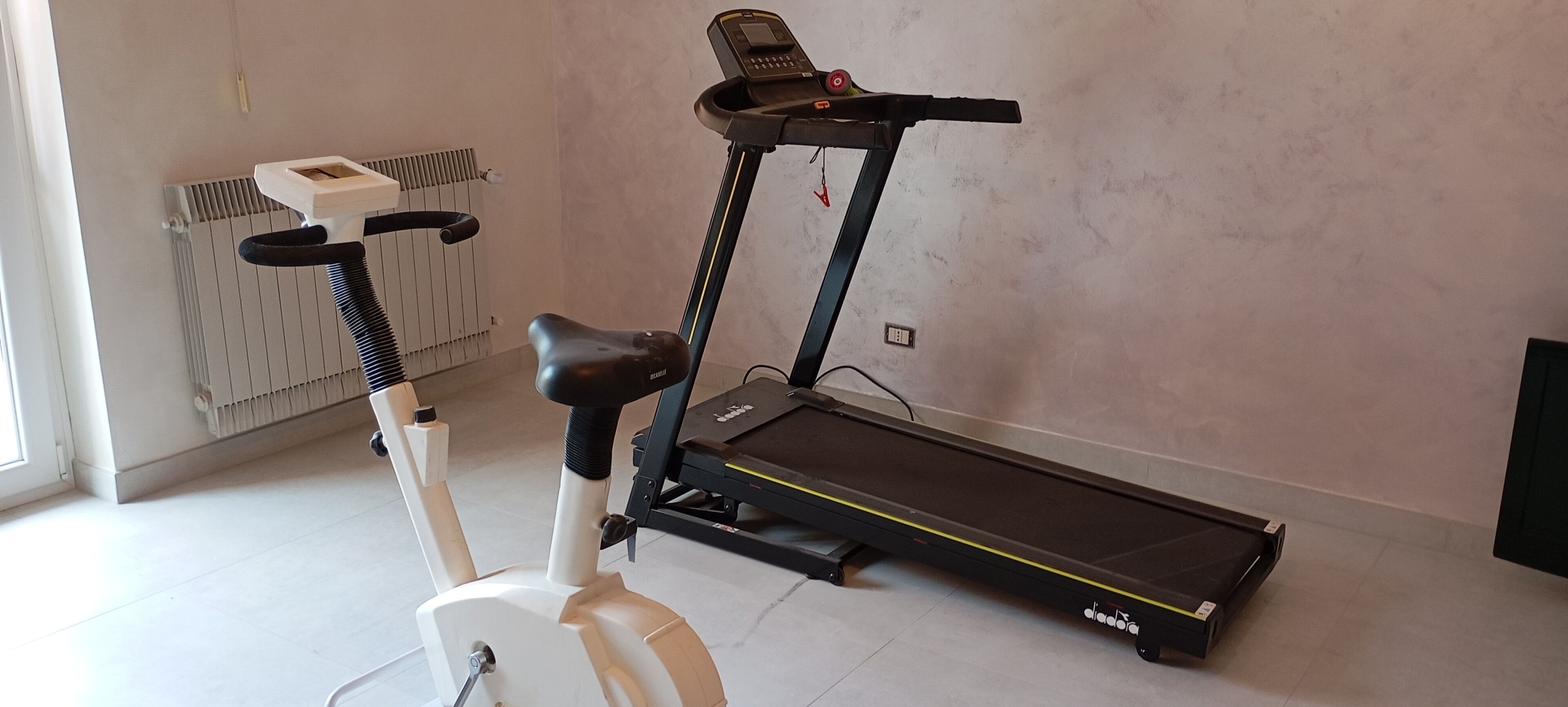 Un vélo d’appartement et un tapis de course se trouvent dans une pièce équipée d’un radiateur et d’une fenêtre.
