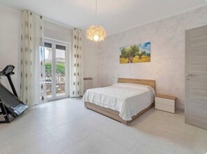 A bedroom with a bed, a treadmill, a sliding glass door, and a painting on the wall.