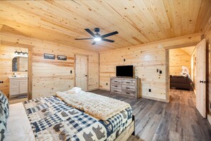 A wooden cabin bedroom with a bed, dresser, TV, and a ceiling fan.