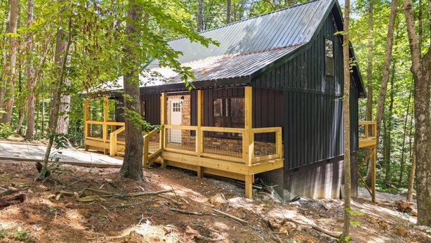 Une cabane dotée d’une terrasse en bois, d’un toit en métal et de fondations en pierre, entourée d’arbres.