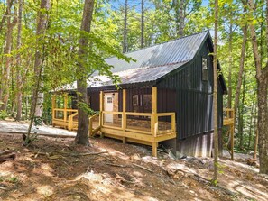 Une cabane dotée d’une terrasse en bois, d’un toit en métal et de fondations en pierre, entourée d’arbres.